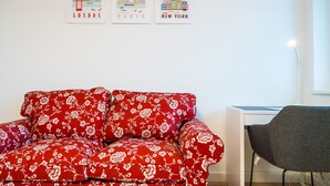 Ein Zimmer mit einem roten, floralen Sofa, einem grauen Sessel und zwei Stadtansichten an der Wand.