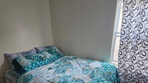 A bedroom with a bed covered in a floral-patterned quilt, a window with patterned curtains, and a plain wall. - Room G Divine Villa and Resorts (Newark)