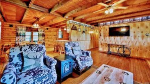 Interior de una cabaña de troncos con techo de madera, un televisor montado en la pared y dos sillones con estampado.