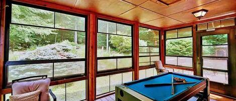 A pool table in a room with large windows overlooking a forested area.