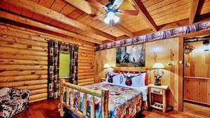 Un dormitorio tipo cabaña de troncos con una cama de madera, un ventilador de techo, un cuadro en la pared y una mesita de noche con lámpara.