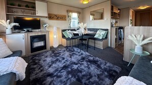 A modern living room with a flat-screen TV, a fireplace, a dining area with a table and chairs, and a cozy seating area with a fur rug. - A Beautiful, Dog Friendly Holiday Home (Mablethorpe)