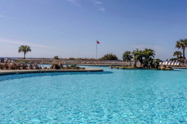 Una gran piscina al aire libre con agua azul cristalina, rodeada de palmeras e izada una bandera.