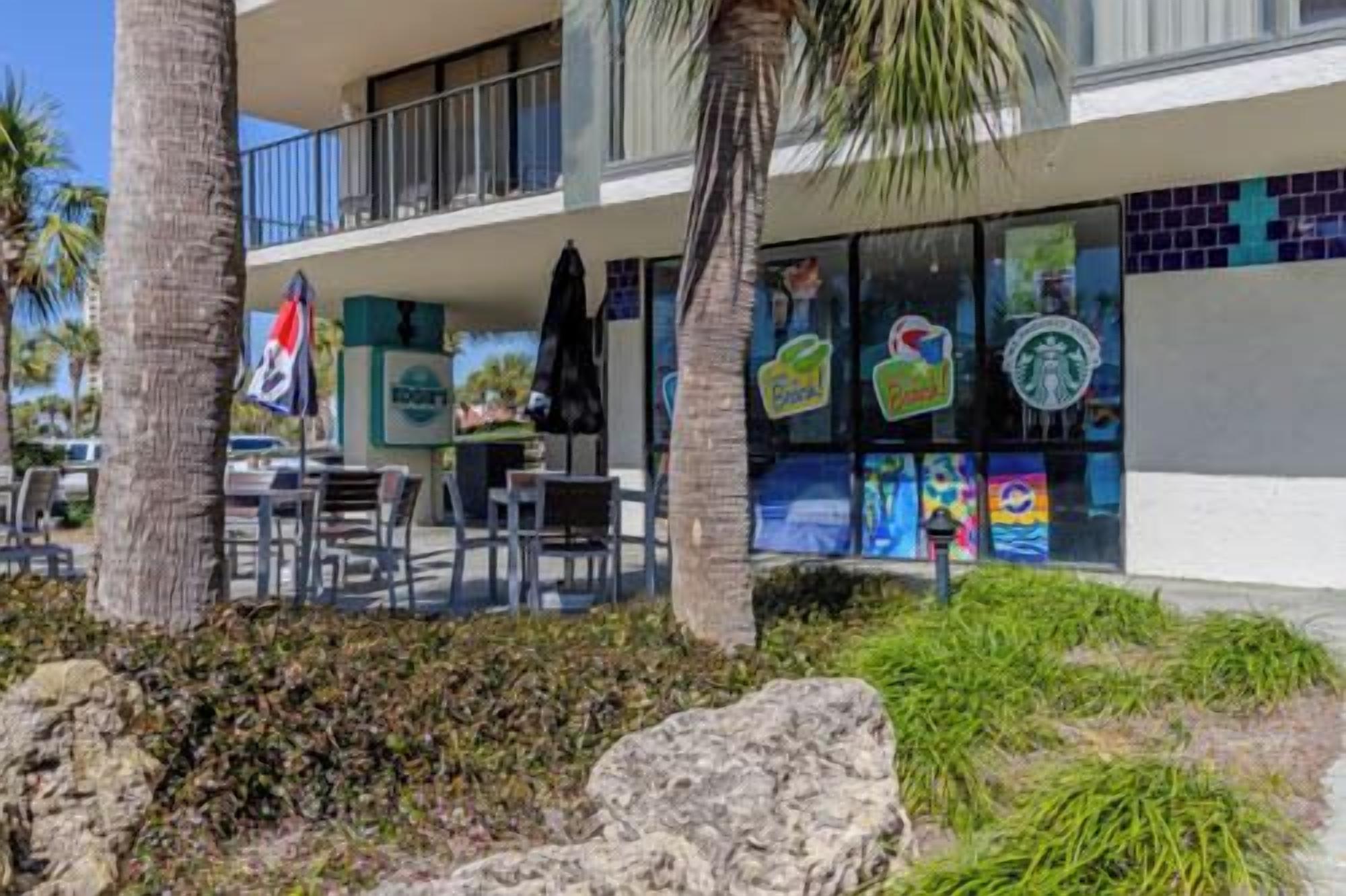An outdoor seating area with tables and chairs, palm trees, and a building with colorful window displays.