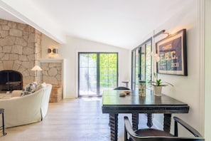 A living room with a stone fireplace, a sofa, a dining table with chairs, and a large window with a view of greenery. - Secluded 3-Bedroom Luxury Retreat near Beach – Perfect Getaway! (Douglas)