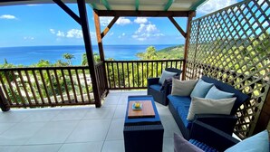 A covered outdoor seating area with a view of the ocean, a wooden table, and a sofa with cushions. - Espace Harmonie vue mer (Deshaies)