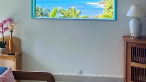 A room with a large window offering a view of the ocean, a wooden side table with a lamp, and a framed picture on the wall. - Espace Harmonie vue mer (Deshaies)