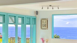 A room with a large window offering a view of the ocean, a framed picture on the wall, and a ceiling light fixture. - Espace Harmonie vue mer (Deshaies)