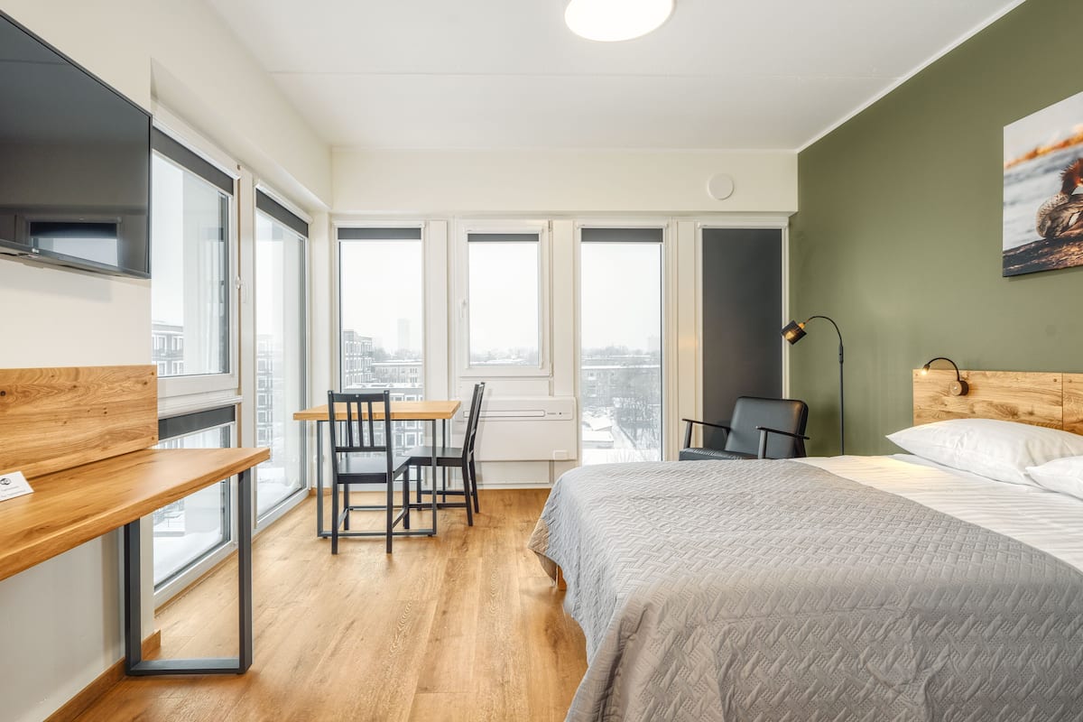 Ein modernes Hotelzimmer mit einem großen Bett, einem Schreibtisch mit Stuhl und Blick auf die Stadt durch große Fenster.