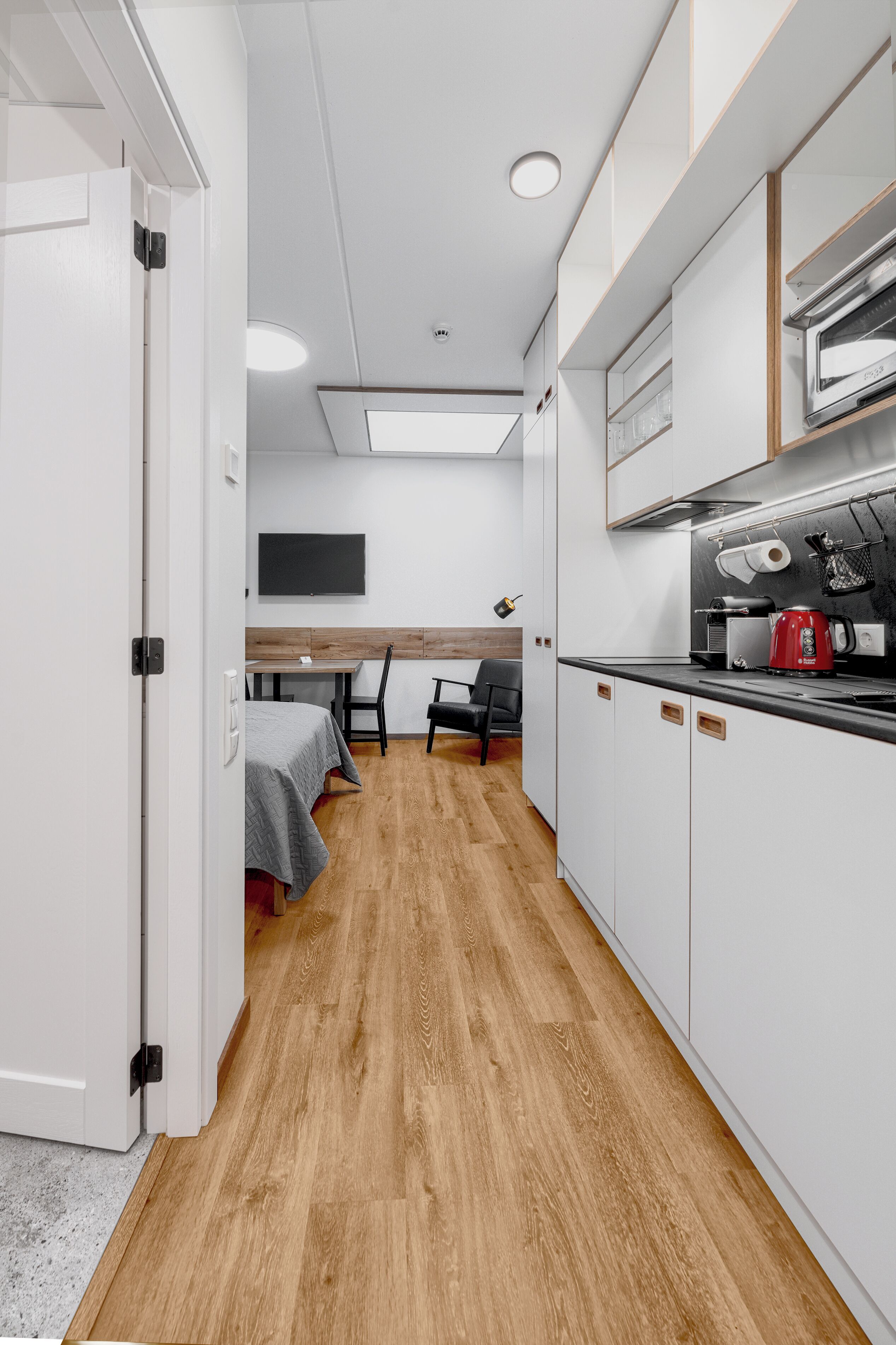 A compact hotel room with a kitchenette, a bed with a gray bedspread, a wooden floor, and a small dining area with a table and chairs.