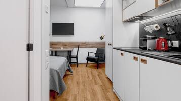 A compact hotel room with a kitchenette, a bed with a gray bedspread, a wooden floor, and a small dining area with a table and chairs.