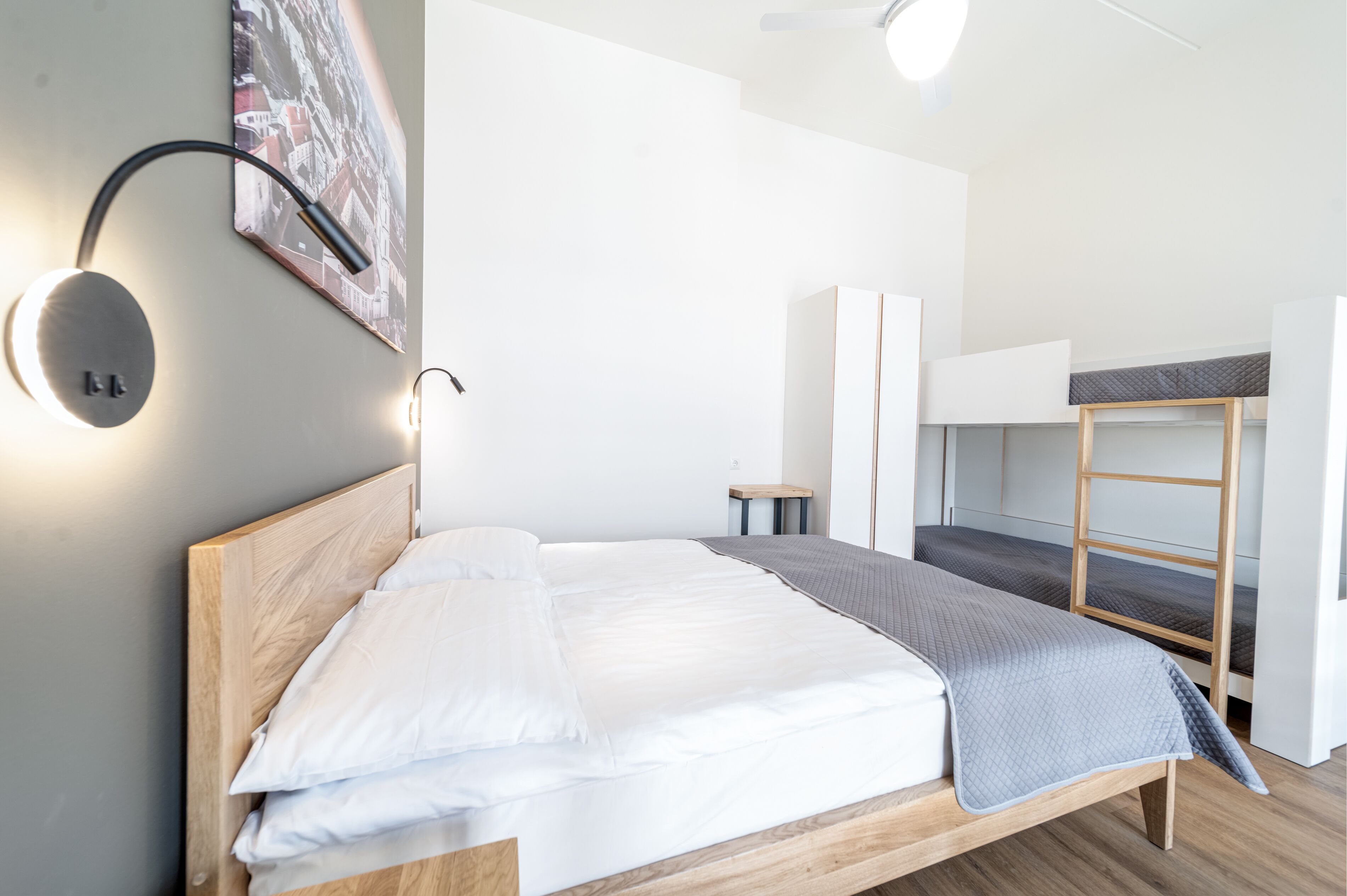 Una habitación de hotel moderna con una cama de madera, una cama literera, una lámpara de pared redonda y una pintura de un paisaje urbano.