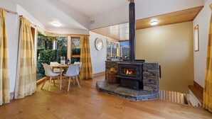 A cozy room with a stone fireplace, wooden flooring, a dining table with chairs, and large windows offering a view of greenery. - Picturesque on Peregrine - close to town with views (Queenstown)