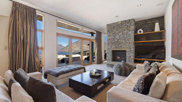 A spacious living room with a stone fireplace, a large TV, and a balcony with a view of the mountains. - Picturesque on Peregrine - close to town with views (Queenstown)