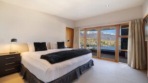 A bedroom with a large bed, a nightstand, a lamp, and a view of a mountainous landscape through a sliding glass door. - Picturesque on Peregrine - close to town with views (Queenstown)