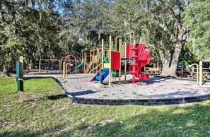 A playground with various play structures including slides and climbing areas, surrounded by trees and a sandy play area.