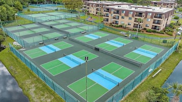 Complejo de canchas de tenis con varias pistas, rodeado por una valla y junto a un edificio residencial.