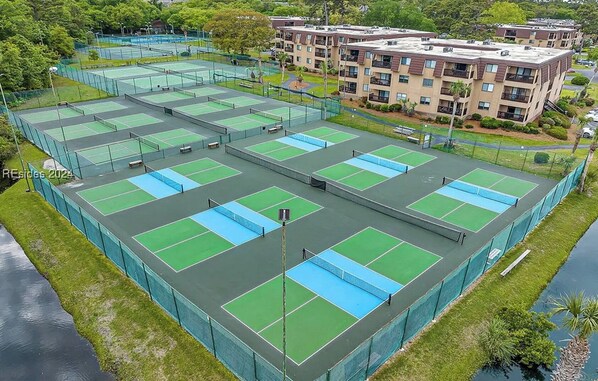 A tennis court complex with multiple courts, surrounded by a fence, and adjacent to a residential building.