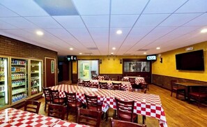 A restaurant with checkered tablecloths, wooden chairs, a brick wall, and a television screen.