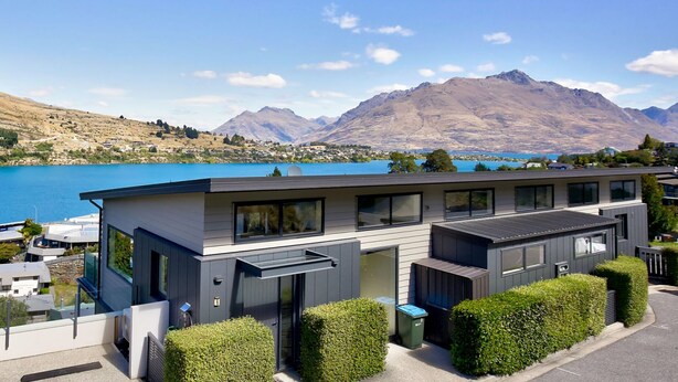 Una casa moderna de dos pisos con techo plano, amplios ventanales y vista a un lago y montañas.