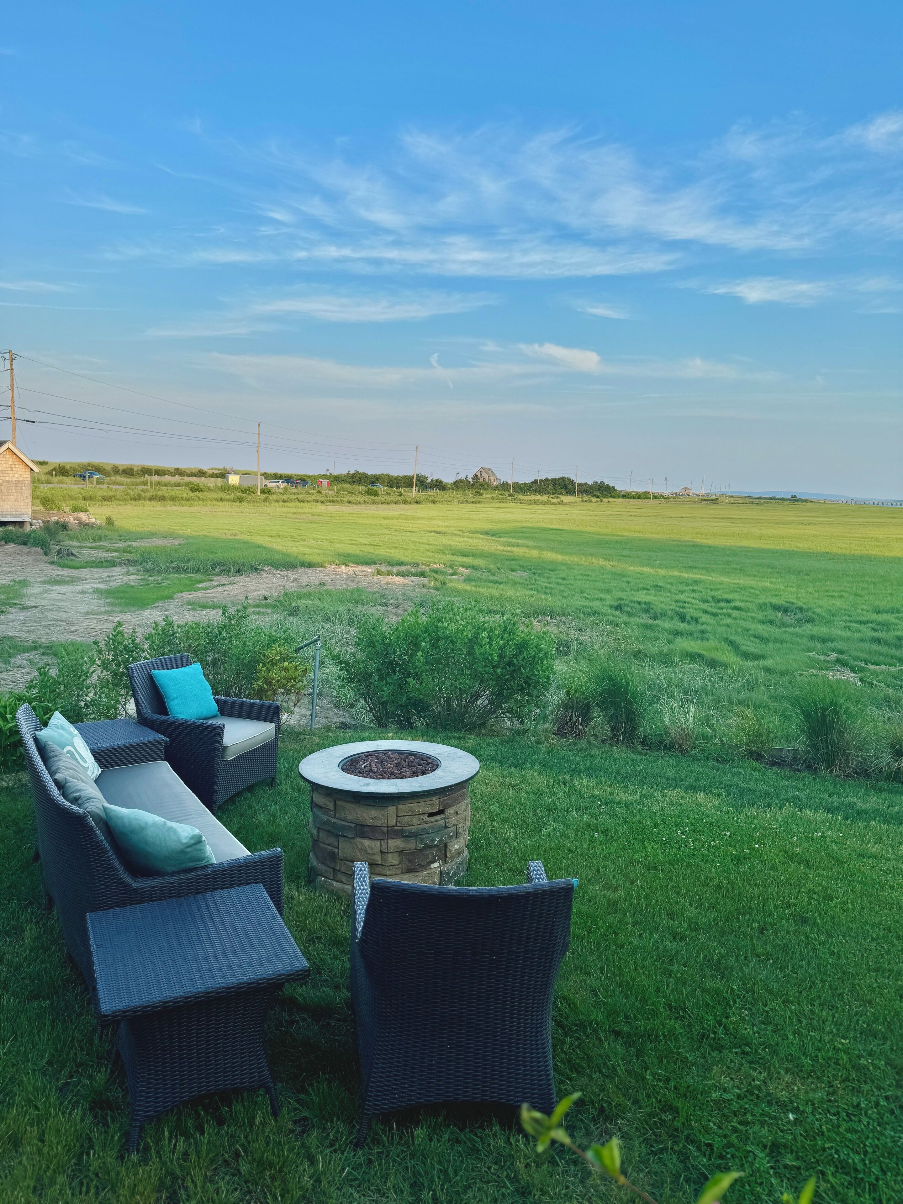 An outdoor seating area with wicker furniture and a stone fire pit, set against a backdrop of a grassy field and a clear sky.