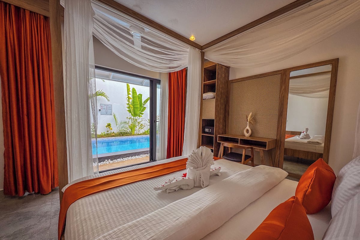 Habitació d'hotel amb un llit gran, una taula de treball, una cadira, un mirall i vista a una piscina exterior.