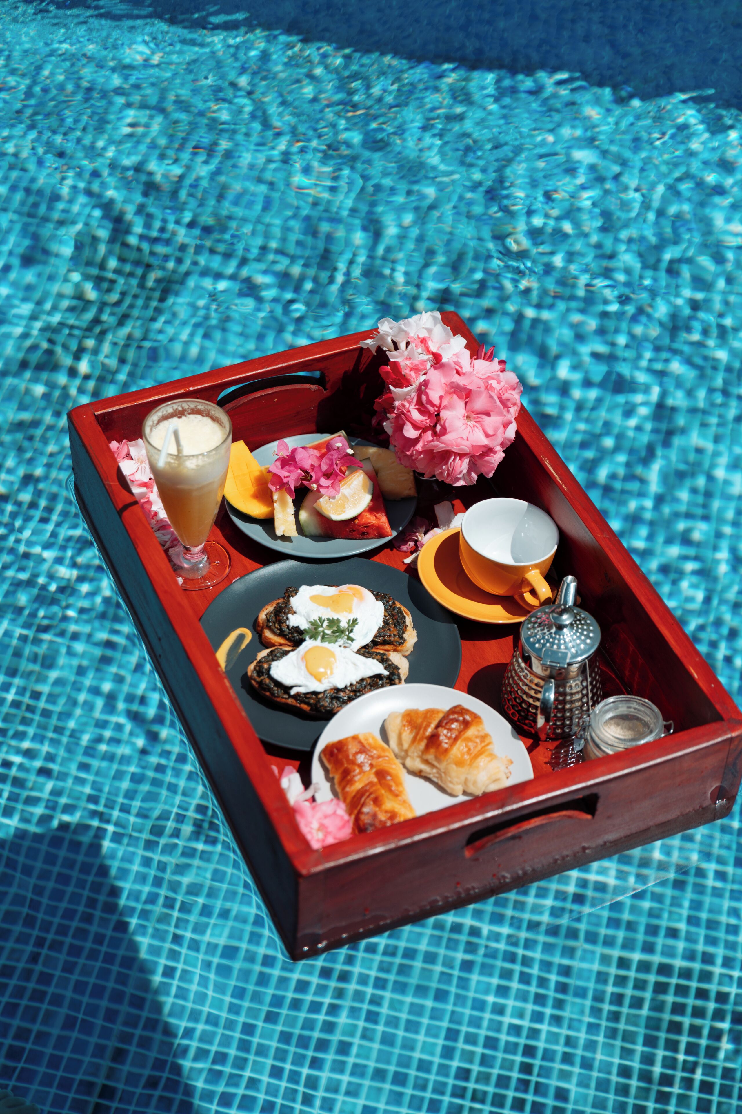 Bandeja con alimentos para el desayuno, como tostadas, huevos, fruta y una bebida, colocada al lado de una piscina.