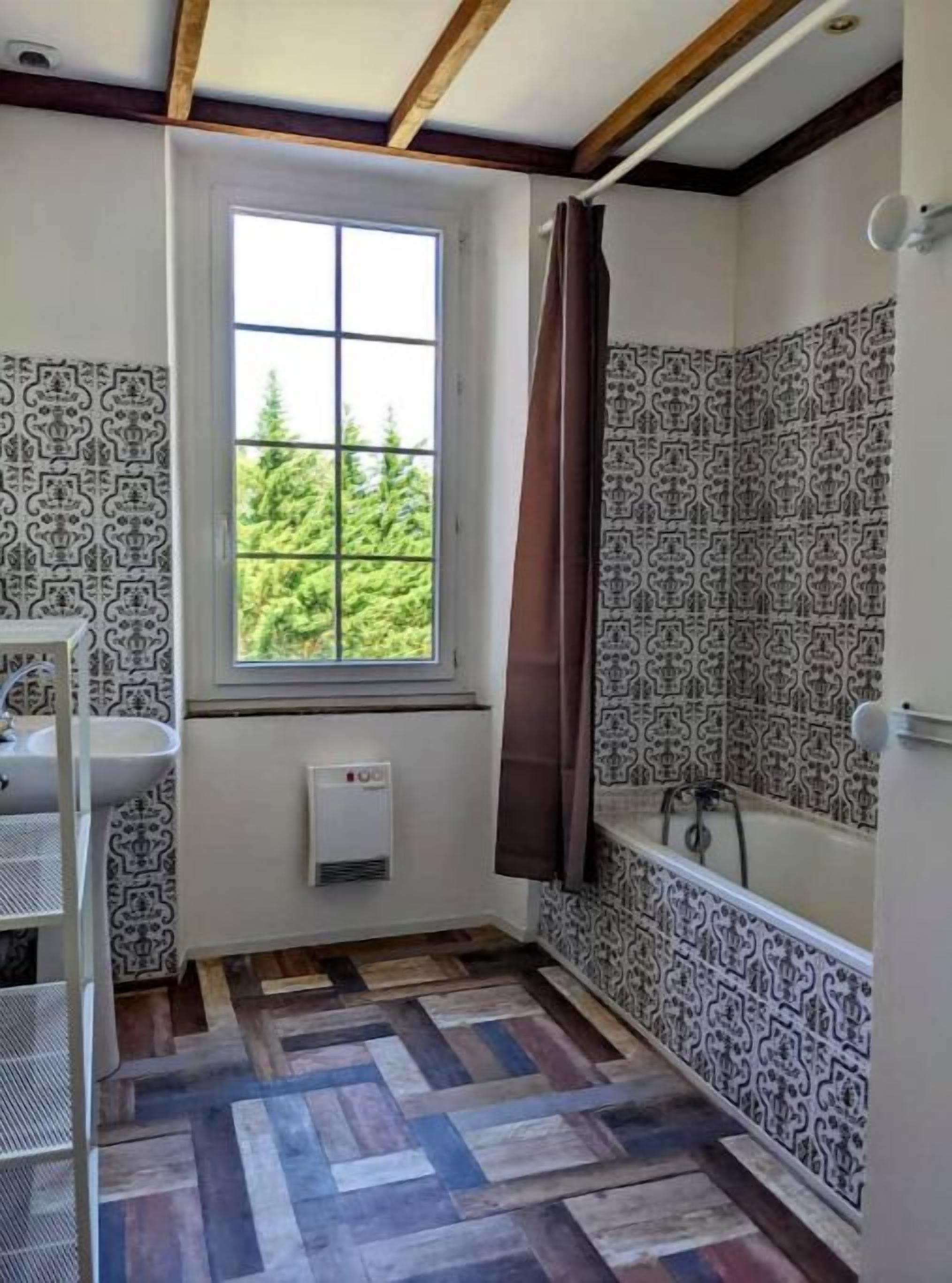 Une salle de bain avec des carreaux décorés, une fenêtre donnant sur un espace verdoyant, une baignoire et un espace douche.