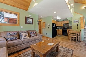 A cozy living room with a sofa, coffee table, and a view of the kitchen. - Fall River Condo • River Views • Near RMNP (Estes Park)