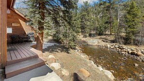 Une cabane en bois dotée d’une terrasse, d’une petite table et de chaises d’extérieur, et offrant une vue dégagée sur un ruisseau bordé de rochers et d’arbres.