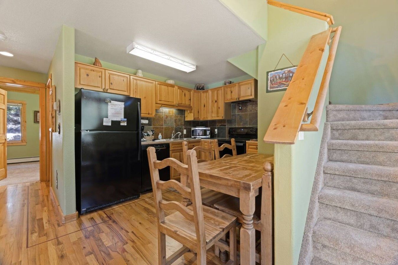 Una cocina con armarios de madera, un refrigerador negro, una mesa de comedor con sillas y una escalera.
