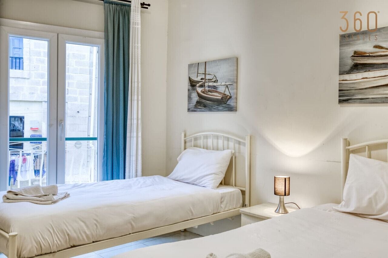Un dormitorio con dos camas, una ventana con cortinas, un cuadro colgado en la pared de un barco y una lámpara de noche.