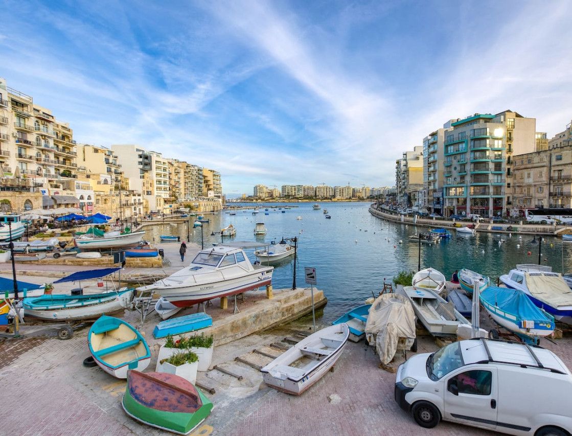 A marina with boats docked, a white van, and buildings along the waterfront.