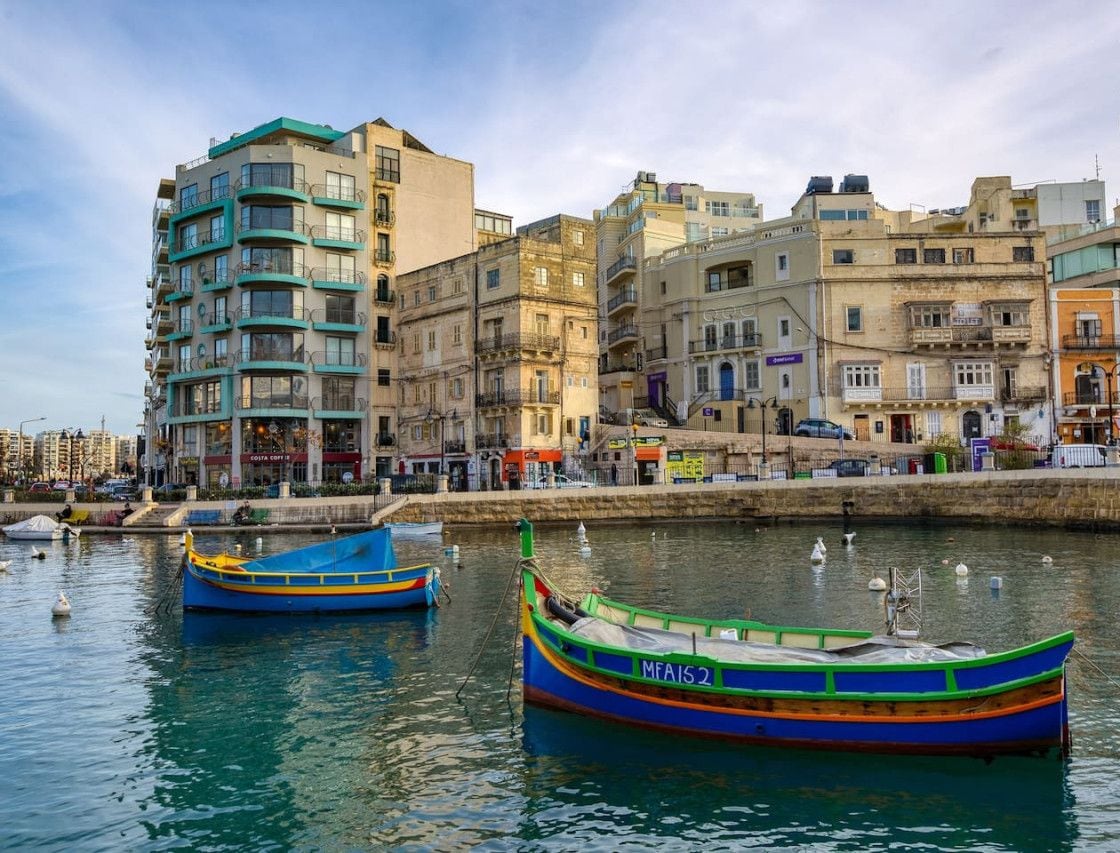 A waterfront with colorful boats docked, a multi-story building with balconies, and a clear sky.