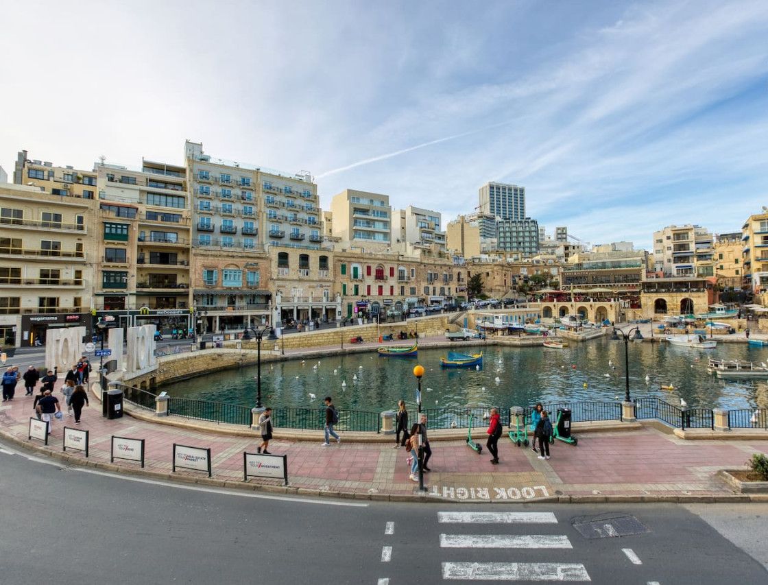 A waterfront promenade with a central water feature, surrounded by multi-story buildings and a clear sky.