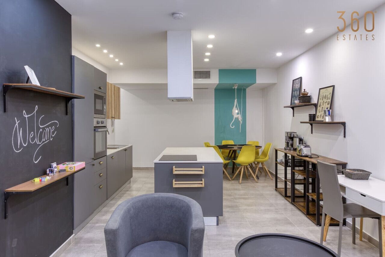 A modern kitchen with a central island, a dining area with yellow chairs, and a 'Welcome' sign on the wall.