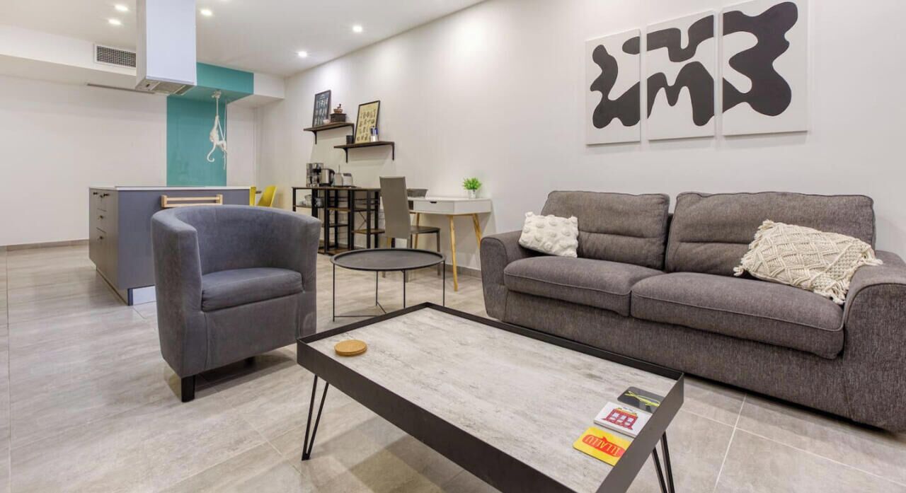 Ein modernes Wohnzimmer mit grauem Sofa, grauem Sessel und Couchtisch. Es gibt eine Küchenzeile mit Esstisch und Stühlen. Der Raum hat ein aufgeräumtes, minimalistisches Design und ein großes abstraktes Kunstwerk an der Wand.