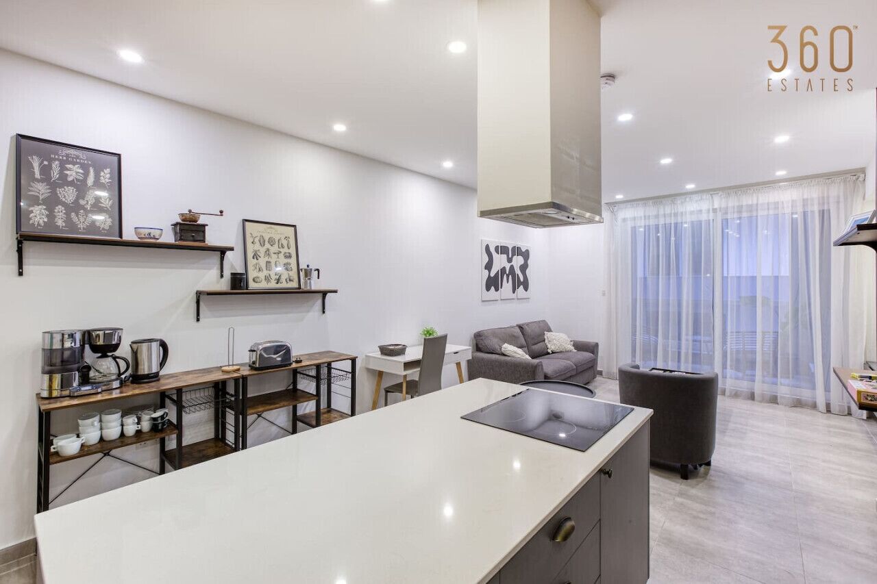 Una cocina moderna con una isla central, un comedor con mesa y sillas, un sofá gris y un amplio ventanal con cortinas translúcidas.