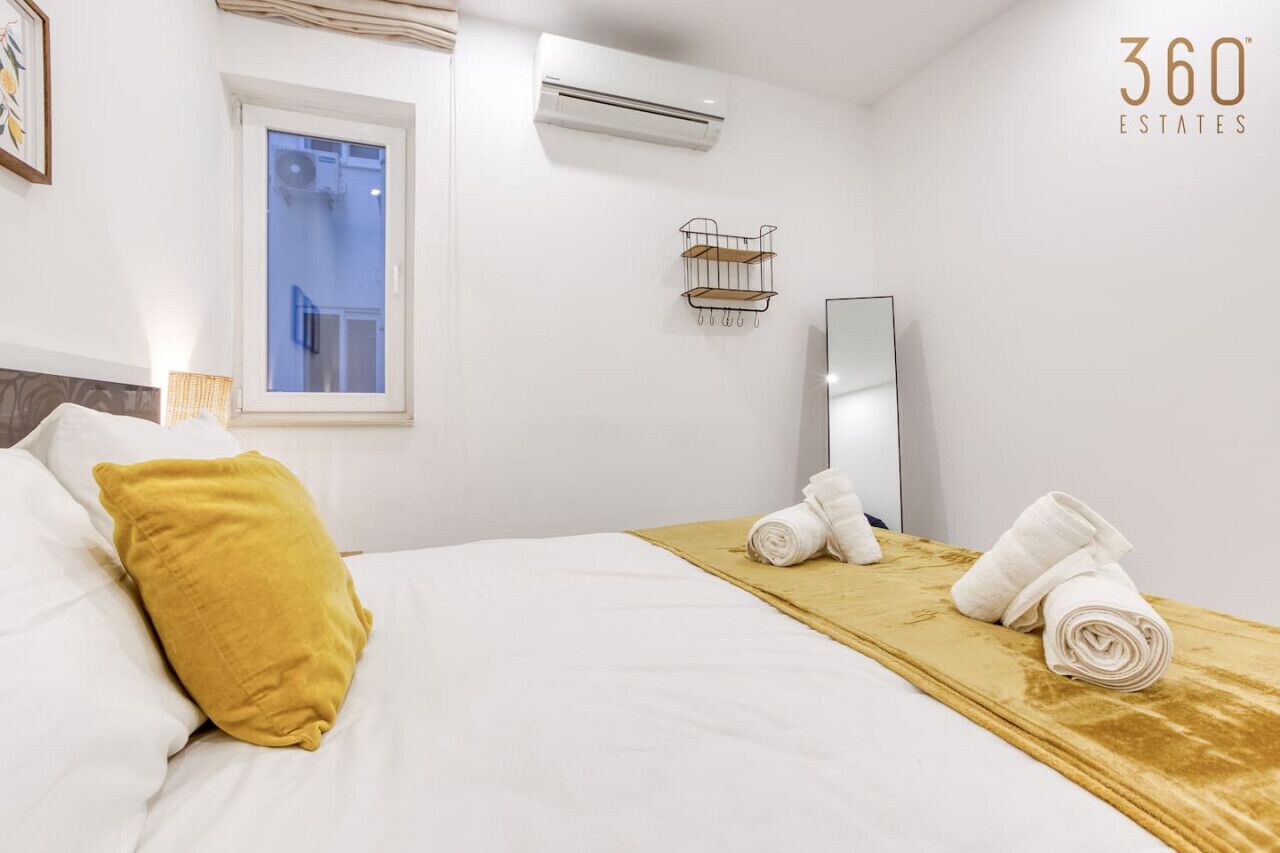 A hotel room with a bed, a window, an air conditioner, a mirror, and a towel rack.