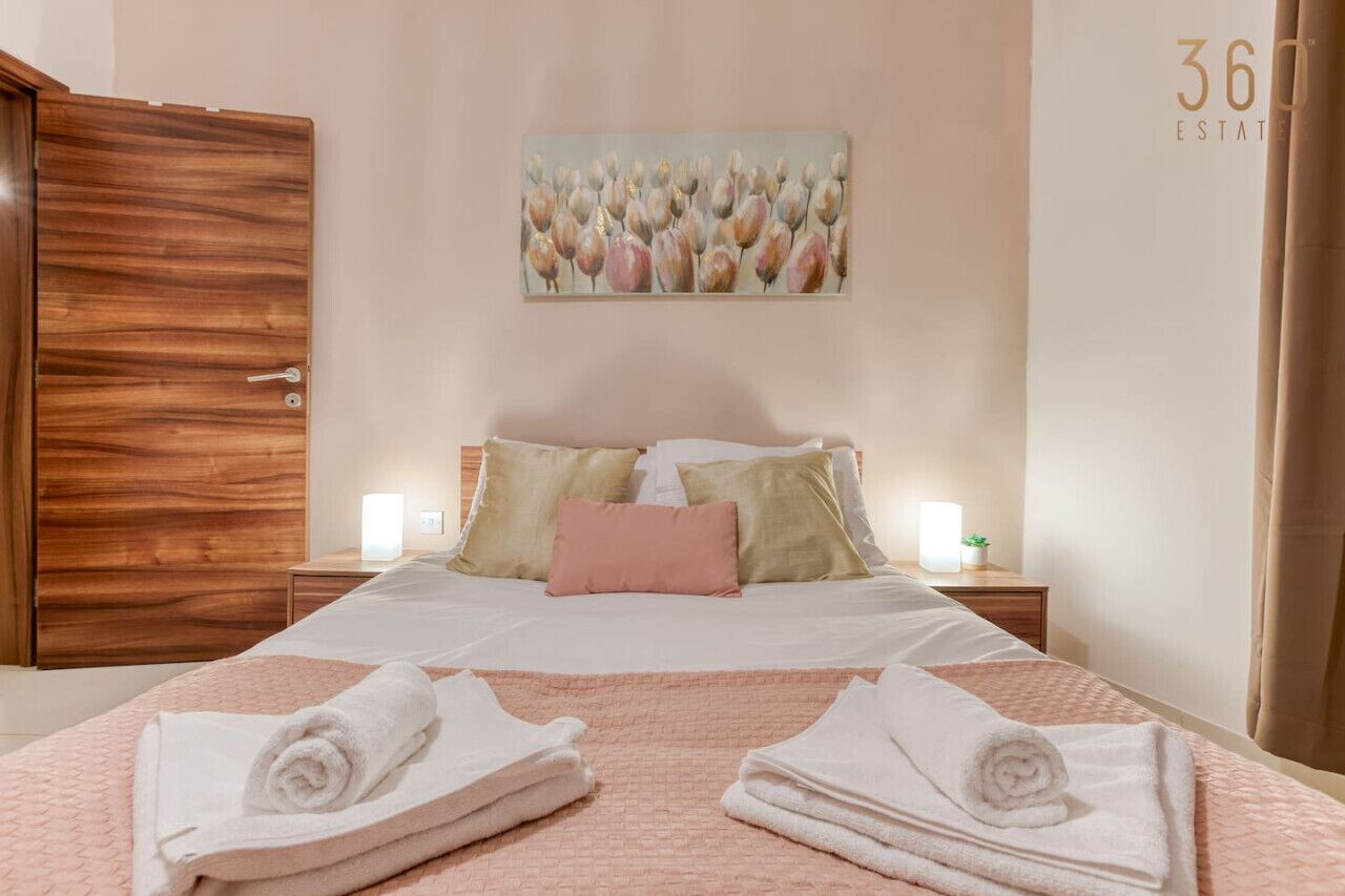 A hotel room with a wooden wardrobe, a bed with white linens, a pink quilt, and a painting of flowers on the wall.