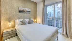 Une chambre avec un grand lit, une table de chevet, une lampe fixée au mur et une fenêtre donnant sur des immeubles.