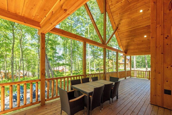 A wooden deck with a table and chairs, surrounded by a wooden railing, overlooking a forested area. - Modern Mountain Getaway w/ Hot Tub & Game Room (McHenry)