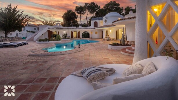 Un área de piscina con una piscina infinita de color blanco, sillones de descanso y una escalera que conduce a un edificio con ventanas arqueadas.