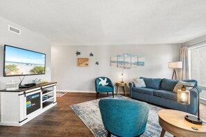 A modern living room with a flat-screen TV, a blue sofa, a teal armchair, and a small side table with a lamp.