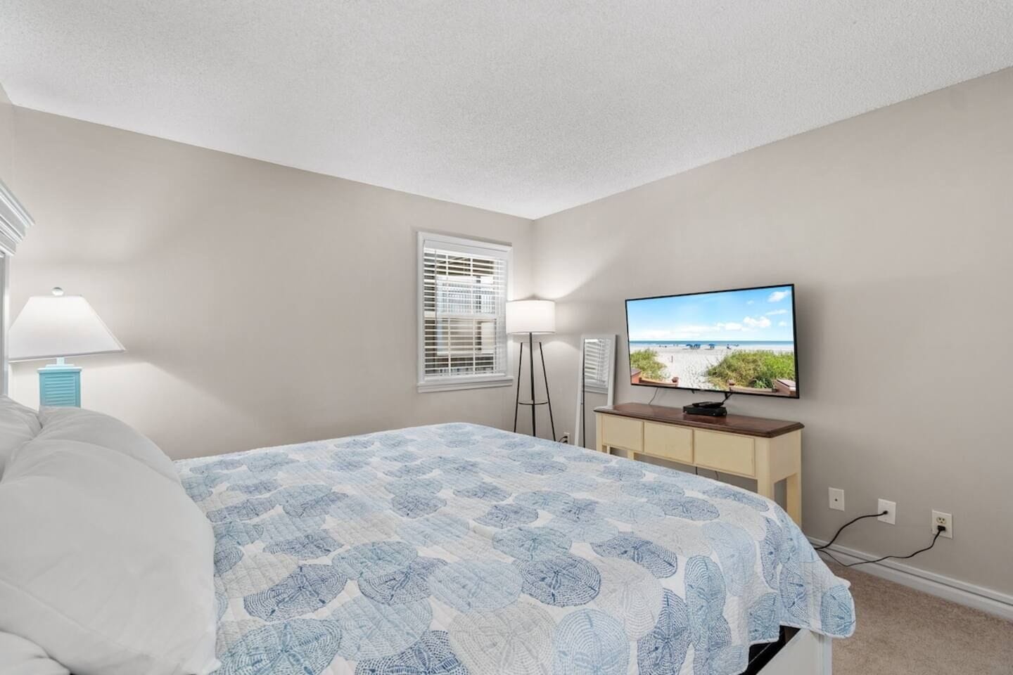 Un dormitorio con cama, televisor sobre un mueble, ventana con persianas y lámpara.