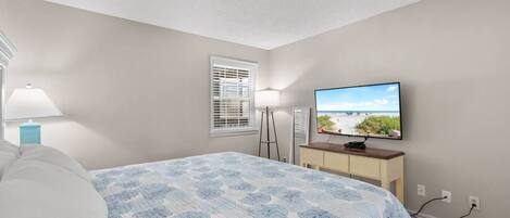 Quarto com cama, televisão em móvel, janela com persianas e abajur.