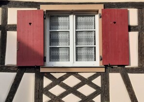 Une fenêtre ornée de rideaux à motifs et de volets rouges, intégrée dans un bâtiment à colombages dont la partie inférieure présente un décor en treillis.