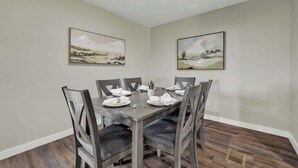 Un comedor con una mesa rectangular dispuesta para seis personas, rodeada por seis sillas con asientos acolchados. La habitación tiene una paleta de colores neutros, con paredes color beige y piso de madera. Dos cuadros de paisajes decoran las paredes sobre la mesa.