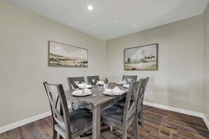 Une salle à manger avec une table rectangulaire dressée pour six personnes, entourée de six chaises dotées d’assises rembourrées. La pièce est décorée dans des tons neutres, avec des murs de couleur beige et un plancher en bois. Deux tableaux représentant des paysages sont accrochés aux murs, au-dessus de la table.