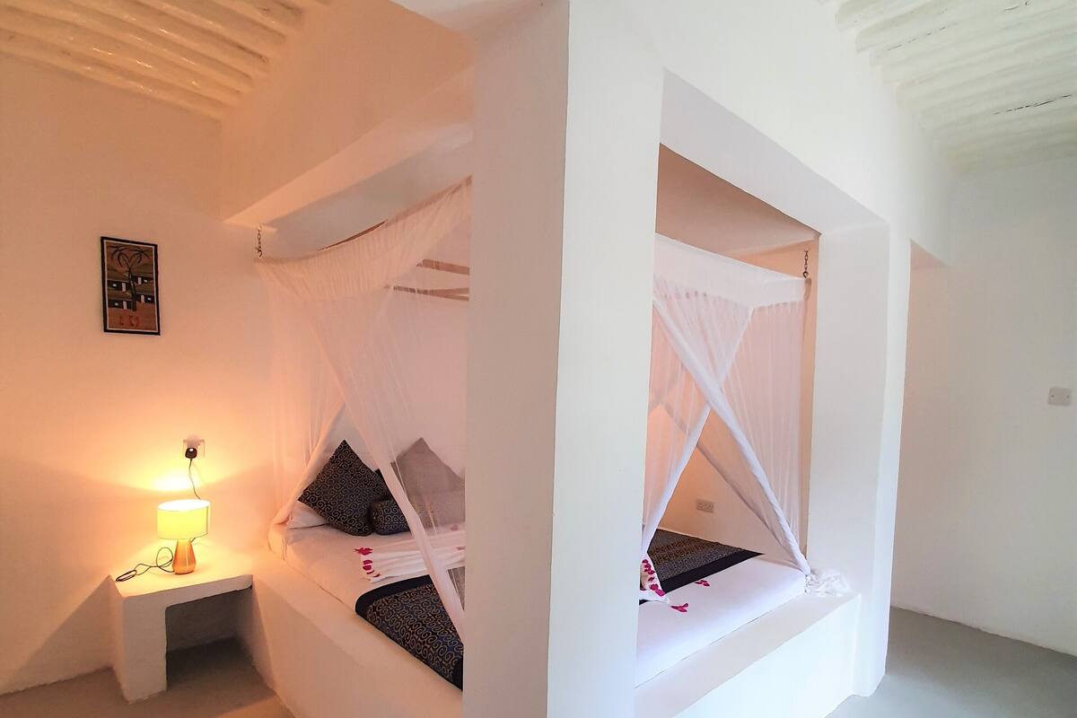 A minimalist bedroom with two beds, each with a canopy, a bedside table with a lamp, and a framed picture on the wall.